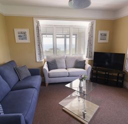 A living room with two sofas a glass coffee table a television on a stand and windows with curtains at Tillicum in Charmouth