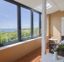 An enclosed porch with large windows overlooking greenery and the sea a wicker sofa with cushions a small table with a bottle and glasses and another table with a vase of flowers at Tillicum in Charmouth