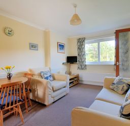 A living room with a wooden dining table and chairs two beige sofas with patterned cushions a small TV on a stand and a window with patterned curtains at Bridge House Apartment in Charmouth