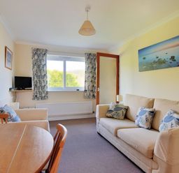 A living room with a beige sofa and armchair with patterned cushions a round wooden dining table chairs a small TV on a wall shelf and a window with curtains at Bridge House Apartment in Charmouth