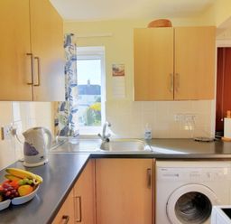 A kitchen with wooden cabinets a kettle a bowl of fruit a washing machine and a window at Bridge House Apartment in Charmouth