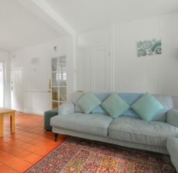 A living room with a light blue sofa with cushions a standing lamp a multicolored rug wooden dining table with benches and a view into a kitchen at Monmouth Cottage in Lyme Regis