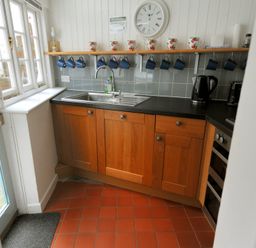 A kitchen corner with wooden cabinets a black countertop a stainless steel sink and mugs hanging under a shelf at Monmouth Cottage in Lyme Regis