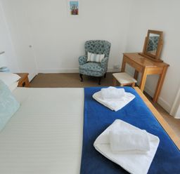 A bedroom with a bed towels a blue patterned chair a wooden dressing table and a framed picture at Monmouth Cottage in Lyme Regis