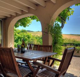 A patio with wooden table and chairs with a bottle and glasses under arches with a view of fields at Willows in Charmouth