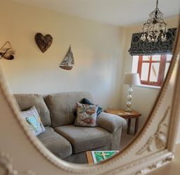 A reflection in a mirror showing a beige couch with patterned cushions near a wooden side table with a lamp and a window with a patterned curtain at 28 Barnes Meadow in Uplyme
