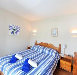 A bedroom with a double bed with blue and white striped bedding two bedside tables with lamps and paintings on the walls at Ivy Cottage in Beer