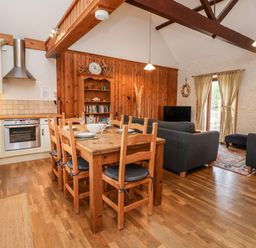A kitchen and living room with a wooden dining table and chairs a bookshelf sofas a television and a kitchen area at 3 Home Farm South in Rousdon