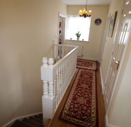 A hallway with a red patterned runner rug on wooden floor a white banister stairs a window with a plant and a chandelier at Vine Cottage in Charmouth