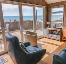 A living room with armchairs a cabinet a window and sliding glass doors showing a deck and ocean view at Ammonite in Lyme Regis
