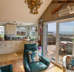 A kitchen with white cabinets and a seating area with green and white armchairs by sliding glass doors leading to a deck with outdoor furniture and a sea view at Ammonite in Lyme Regis