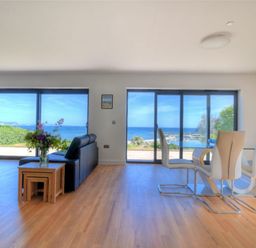 A living room with black sofas and a dining table with white chairs by large windows overlooking the sea at Harbour Heights Cottage in Lyme Regis