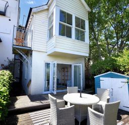 An outdoor patio area with a round table and four chairs next to a blue and white shed and a two-story house with white siding and large windows at Little Bay View in Lyme Regis