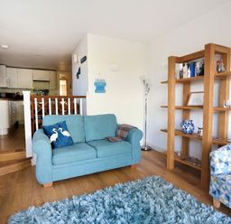 A living room with blue sofa, patterned armchair, wooden shelving unit, and kitchen area at Little Bay View in Lyme Regis