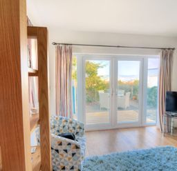 A living room with a patterned armchair a wooden shelf a television on a glass stand and glass doors leading to an outdoor patio at Little Bay View in Lyme Regis