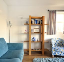 A living room with a blue sofa a patterned armchair a wooden bookshelf and a window with striped curtains at Little Bay View in Lyme Regis