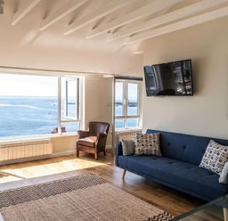 A living room with a blue sofa pillows two brown armchairs a rug and large windows overlooking the sea at 3 Beach Way House Lyme Regis