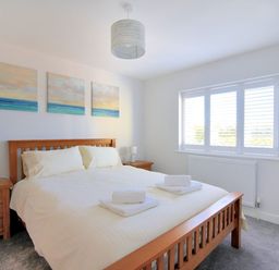 A bedroom with a wooden bed dressed in white linens two bedside tables with lamps three paintings above the bed a window with white shutters and a small TV on a stand at Shire View in Lyme Regis