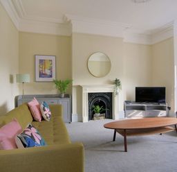 A living room with a green sofa pink and floral cushions a wooden coffee table a fireplace with plants and a round mirror at 7 Seafield Road in Seaton