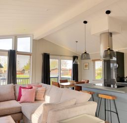 An open living room and kitchen area with a beige sectional sofa with pink cushions a kitchen island with stools pendant lights and large windows at Hazelwood 4 @ Pinewood Retreat in Pinewood