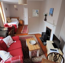 A living room with a red sofa white pillows a wooden coffee table a rug a fireplace a TV and a single bed in the background at Driftwood in Lyme Regis
