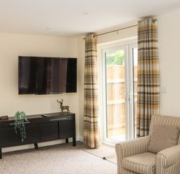 A living room with a wall-mounted TV a black cabinet a striped armchair and a glass door with plaid curtains at Sawtons Cottage 2 in Dawlish