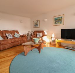 A living room with brown sofas a glass coffee table a blue round rug a television on a wooden stand and framed paintings on the walls at Port View in Falmouth