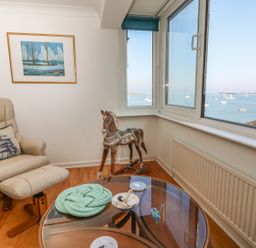 A room with a beige recliner chair and footrest a wooden rocking horse a glass round coffee table and a window showing a sea view at Port View in Falmouth