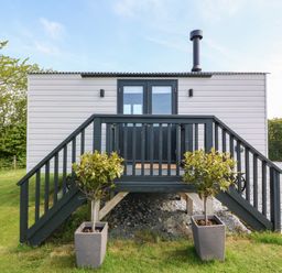A small elevated cabin with black railings and stairs flanked by two potted plants on a grass lawn at Cwt Y Rhos in Valley