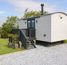 A small white wagon with a black chimney and black stairs on a gravel driveway surrounded by grass and bushes at Cwt Y Rhos in Valley