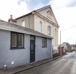 A street with a small gray house with black door and windows next to a large ornate building and other buildings in the background at Ty Capel Bach in Rachub