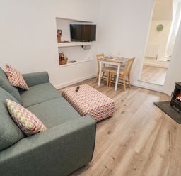 A living room with a green sofa, patterned ottoman, small dining table, wall-mounted TV, and wood burning stove at Ty Capel Bach in Rachub