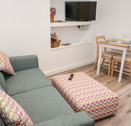 A living room with a green sofa and matching cushions next to a patterned ottoman a wall-mounted TV and a small dining table with two chairs at Ty Capel Bach in Rachub