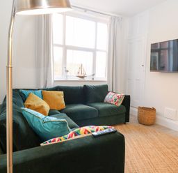 A living room with a green sectional sofa with multicolored cushions a wall-mounted television a floor lamp and a woven basket at Flat 1 in Hunstanton