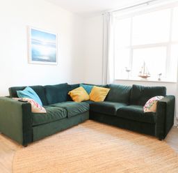 A living room with a green corner sofa yellow and blue cushions a floor lamp a round rug and a window with decorations at Flat 1 Hunstanton