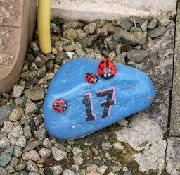 A blue painted rock with number 17 and ladybug decorations on gravel in an outdoor area