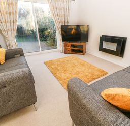 A living room with two grey sofas and yellow cushions a TV on a wooden stand a wall-mounted fireplace and sliding glass doors with curtains at Dunes View in Cubert