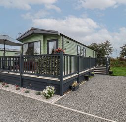 A green mobile home with a black wooden deck and railing surrounded by gravel and flowers at Jean's Place in East Heslerton near West Heslerton