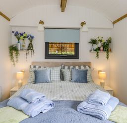 A bedroom with a double bed side tables lamps folded towels flowers on shelves and a small window at High Rigg Shepherd's Sunrise in Brampton Cumbria