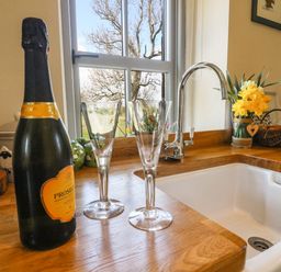 A kitchen countertop with a bottle of prosecco and two glasses next to a sink and a window at Herdie Hideaway in Appleby-In-Westmorland