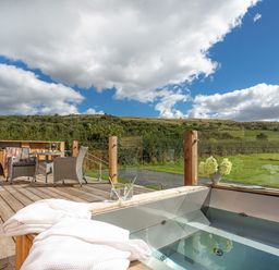 An outdoor wooden deck with a hot tub towels table chairs barbecue and grassy hills in the background at Little Heaning in Cartmel