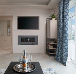 A living room with a wall-mounted TV and fireplace shelves with books and a plant and a bottle with glasses on a table near window with blue patterned curtains at Cartref in Benllech