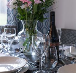 A dining table set with plates glasses cutlery a wine bottle and a vase with flowers at Cartref in Benllech
