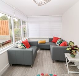 A seating area with gray sofas and colorful cushions by windows with white blinds and a white side table with a plant at Daresfield in Cemaes Bay