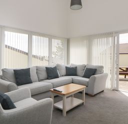 A sitting room with a grey corner sofa and armchair with cushions a wooden coffee table and vertical blinds on windows at Harcombe House Bungalow 11 in Chudleigh