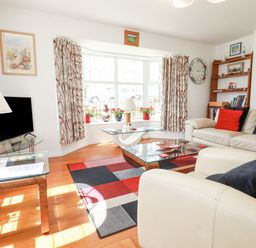 A living room with white sofas and red and black pillows a glass coffee table a patterned rug bay window with plants and patterned curtains at 2 Old Talbot Cottages Lostwithiel