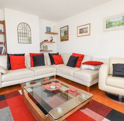 A living room with a beige leather sofa and armchair with red and black pillows at 2 Old Talbot Cottages in Lostwithiel