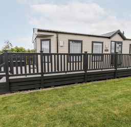 An exterior view of a beige mobile home with black framed windows and black railing on a deck with grass in the foreground at Lazy Daze in Carnaby near Bridlington