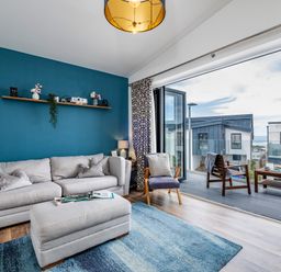 A living room with a grey sofa and ottoman next to a glass door leading to a balcony with chairs at Beach Haven in Perranporth