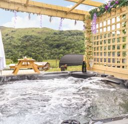 An outdoor hot tub under a wooden pergola with hanging flowers and a bottle with glasses next to it and a picnic table in the background at Hartsop Magic - Crossgate Luxury Glamping in Hartsop near Glenridding